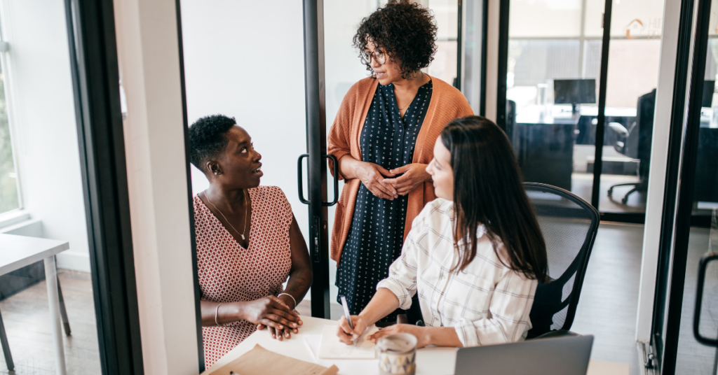 Three colleagues having a collaborative discussion in a glass-walled office, highlighting teamwork, communication, and inclusivity.