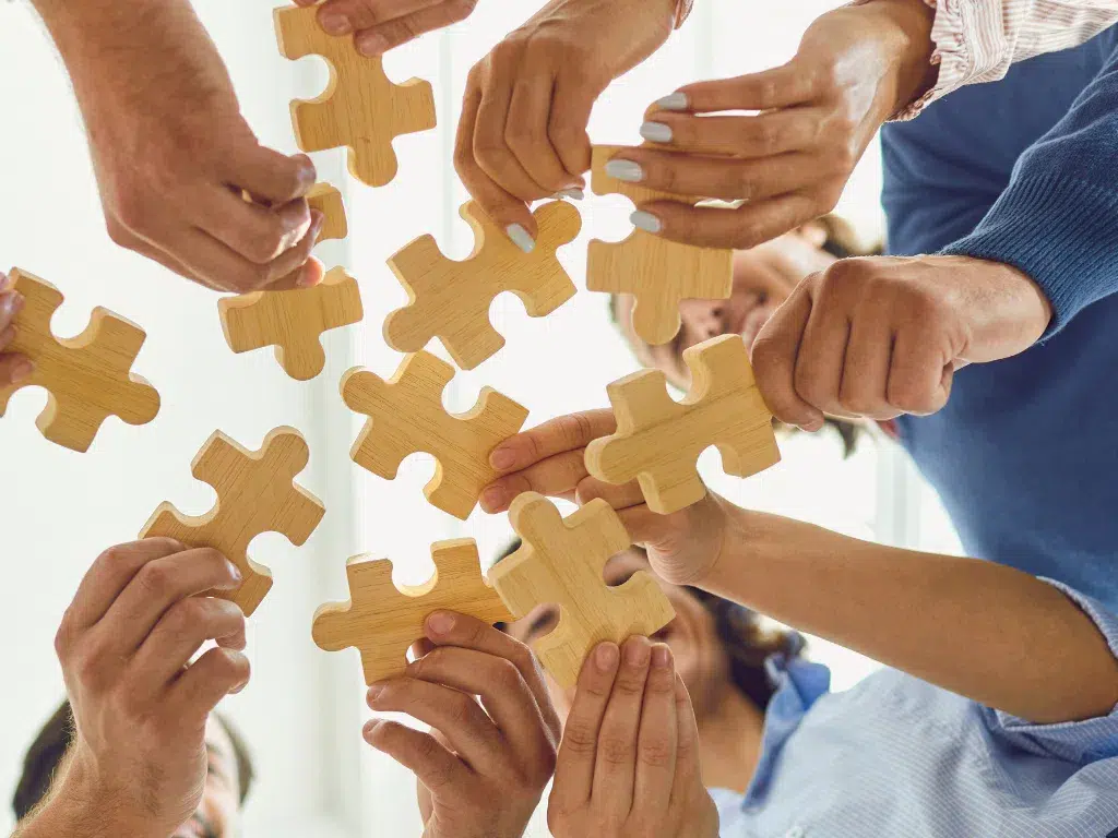 A group of hands holding wooden puzzle pieces, coming together to assemble them, symbolizing teamwork, collaboration, and solving problems collectively.